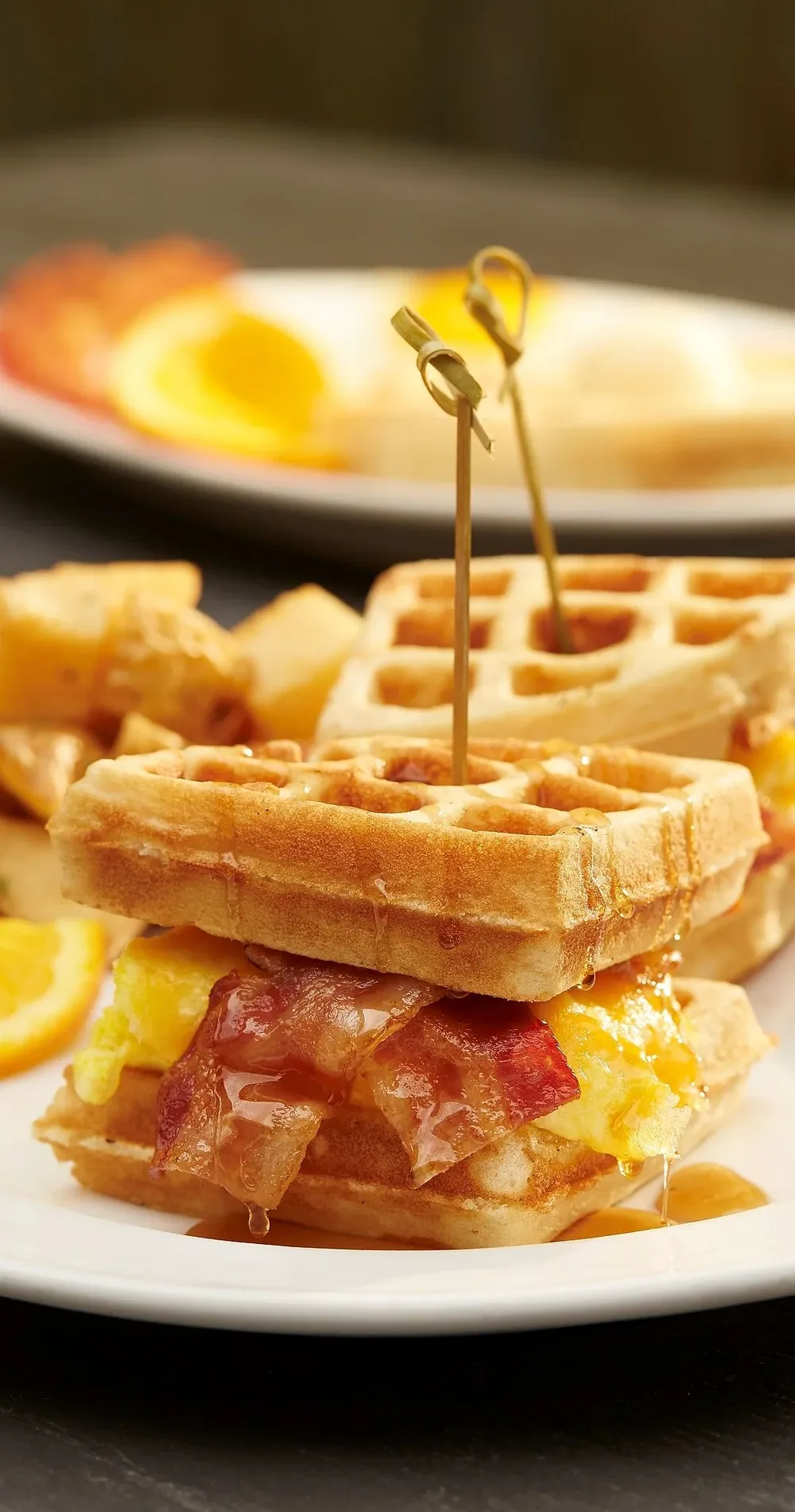 A waffle breakfast sandwich with bacon, egg and cheese, served with potatoes and orange